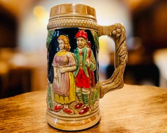 Puede incluir: Una jarra de cerveza de cerámica con un diseño en relieve detallado. La jarra presenta una pareja con vestimenta tradicional, con un hombre con abrigo rojo y una mujer con vestido. El asa tiene forma de rama de árbol y los colores son tonos tierra.
