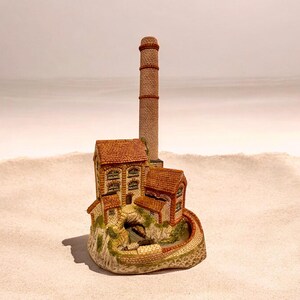 May include: A detailed miniature model of a brick factory with a tall chimney. The buildings have red tile roofs and arched windows. The model sits on a textured base with a winding brick wall and greenery.