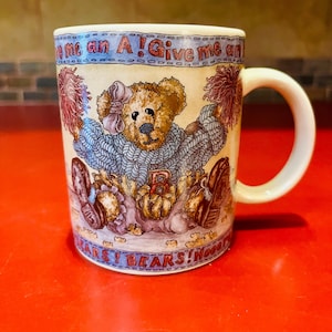 May include: A ceramic mug with a cream-colored background features a cartoon teddy bear wearing a blue sweater and holding pom-poms. The mug has a white handle and the text "Give me an A! Bears! Hooray!" in blue.