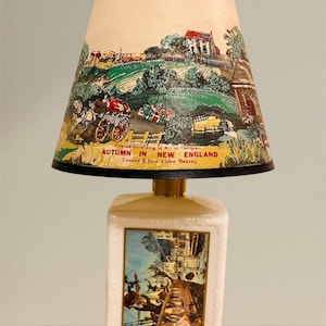 May include: A vintage table lamp with a cream-colored rectangular base and a conical lampshade. The lampshade features a colorful illustration of a New England autumn scene with text that reads "Autumn in New England".