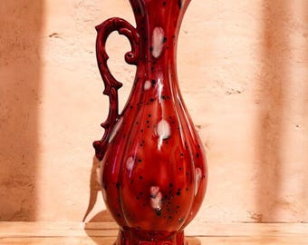 May include: A tall, red ceramic pitcher with a glossy finish. The pitcher features a decorative handle and a flared rim. White and black speckles dot the surface. The pitcher sits on a decorative base, and is a decorative home accent.
