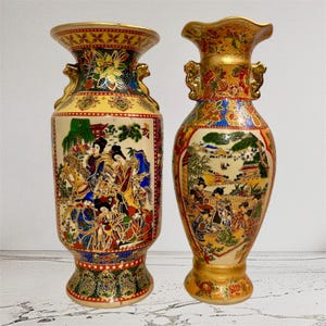 Op de afbeelding: Twee keramische vazen met versierde gouden randen en handvatten. De linker vaas heeft een crèmekleurig paneel met figuren te paard en bloemmotieven. De rechter vaas heeft een geschulpte rand en een gedetailleerd landschap met figuren.