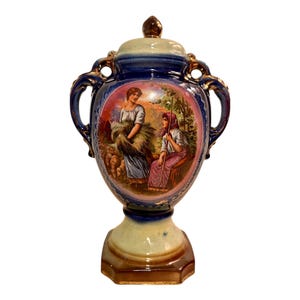 May include: A vintage ceramic urn with a lid, featuring a hand-painted scene of two women in a pastoral setting. The urn has a blue and gold color scheme, with a pedestal base and two ornate handles. The lid is topped with a brown finial.