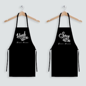 May include: Two black aprons with white text and graphics. The apron on the left says "Head Chef" with a chef's knife and spatula. The apron on the right says "Sous Chef" with a chef's knife, fork, and spatula. Both aprons have the text "Your Name" below the graphic.