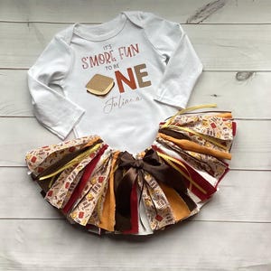 May include: A white long-sleeve bodysuit with the text IT'S S'MORE FUN TO BE ONE and a s'more graphic, paired with a multi-colored tutu skirt featuring brown, gold, and patterned fabric strips tied with a dark brown satin ribbon bow at the center.