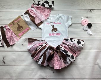 May include: A rodeo-themed baby outfit set, including a white onesie with "My 1st Rodeo" and a bucking bronco graphic, a tutu skirt, a headband, and a banner. The set features pink, brown, white, and cow print accents.