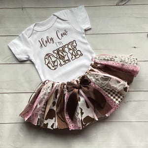 May include: A white baby bodysuit with the text "Holy Cow I'm ONE" in brown lettering, paired with a multi-layered tutu skirt. The skirt features brown, pink, and white fabrics, including cow print and lace, and a large brown bow.