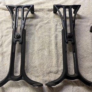 May include: Two black cast iron table legs with an ornate, open-work structural design and a central adjustable mechanism. A single black adjustable wrench is positioned to the right of the legs, providing a sense of scale for the metal components.