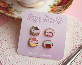 Puede incluir: Un conjunto de cuatro pines de esmalte con pequeños dulces, que incluyen un pastel de arándanos, un pastel de fresa, un mochi rosa y un mochi verde. Los pines están en una tarjeta morada con el texto "tiny treats by donatsuya".