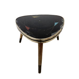 May include: A triangular side table with a black top featuring thin white curved lines and small red, yellow, and blue geometric accents. The table has a metallic gold-toned rim and three tapered wooden legs with dark brown painted tips.
