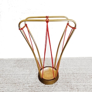 Puede incluir: Un paragüero dorado con un diseño geométrico. El soporte presenta una base circular y una serie de varillas metálicas curvas conectadas por cuerdas rojas. La parte superior tiene forma rectangular. El interior es de color beige claro.