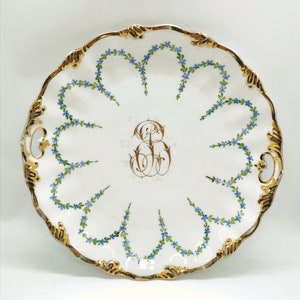 Puede incluir: Un plato de servir de porcelana blanca con un borde dorado y un diseño floral. El plato tiene dos asas y un monograma en el centro.
