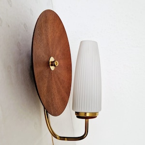 Puede incluir: Una lámpara de pared de estilo modernista de mediados de siglo con una placa trasera circular de madera y una pantalla de vidrio acanalada blanca.