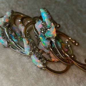 May include: An ornate brooch featuring iridescent opal stones and silver and gold-toned metalwork. The opal stones display a range of colors, including blue, green, and pink. The brooch has a complex, layered design with curved elements.