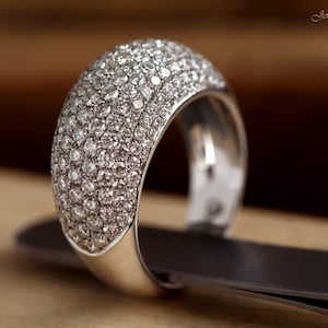 May include: A wide, silver ring encrusted with numerous small, sparkling diamonds. The ring is set on a dark surface, with a blurred background. The ring's band is thick and polished, reflecting light. The jewelry is from Jewellery by Spektor.