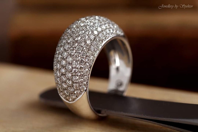 May include: A wide, silver ring encrusted with numerous small, sparkling diamonds. The ring is set on a dark surface, with a blurred background. The ring's band is thick and polished, reflecting light. The jewelry is from Jewellery by Spektor.
