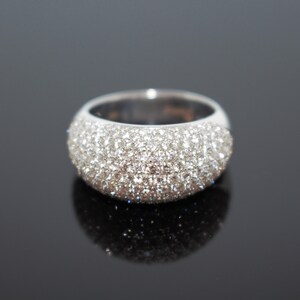 May include: A wide, silver-toned ring with a domed surface, completely encrusted with small, sparkling, clear stones. The ring sits on a reflective black surface, creating a mirrored effect. The ring's design is simple yet elegant.
