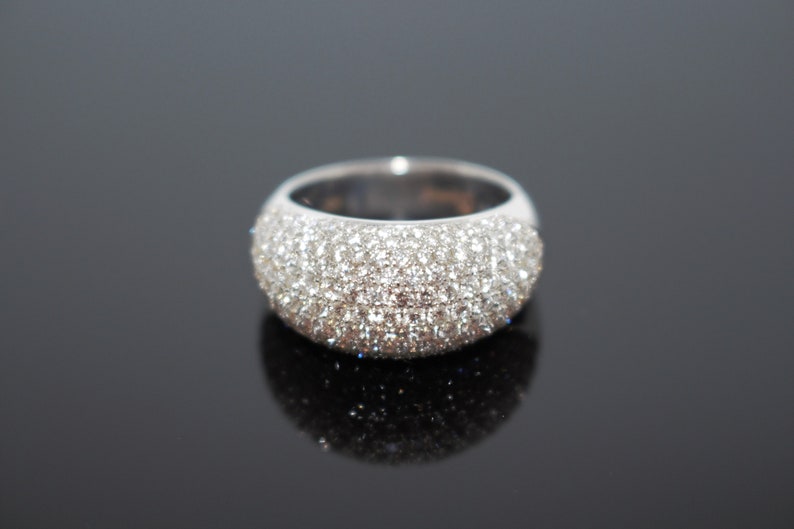 May include: A wide, silver-toned ring with a domed surface, completely encrusted with small, sparkling, clear stones. The ring sits on a reflective black surface, creating a mirrored effect. The ring's design is simple yet elegant.
