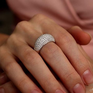 May include: A silver ring with a domed surface, completely covered in small, sparkling clear stones. The ring is on a person's finger, with the hand and arm visible. The jewelry is set against a soft pink background.