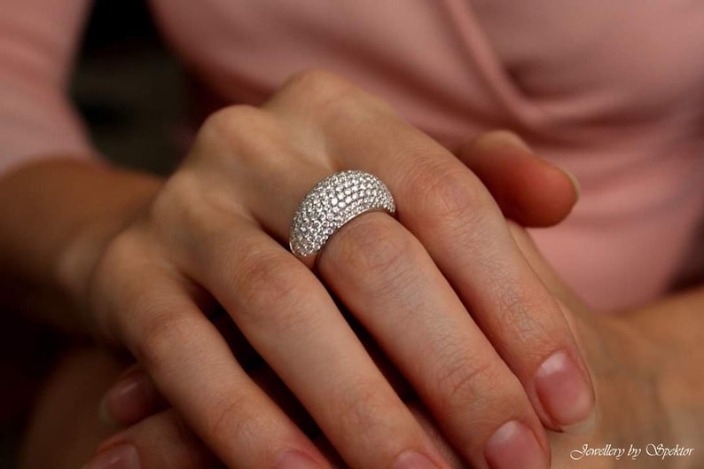 May include: A silver ring with a domed surface, completely covered in small, sparkling clear stones. The ring is on a person's finger, with the hand and arm visible. The jewelry is set against a soft pink background.