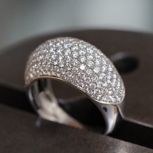 May include: A close-up of a silver ring with a domed top fully encrusted with small, sparkling clear gemstones. The ring is set on a dark, metallic surface, highlighting its brilliance.