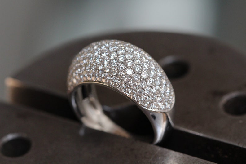 May include: A close-up of a silver ring with a domed top fully encrusted with small, sparkling clear gemstones. The ring is set on a dark, metallic surface, highlighting its brilliance.