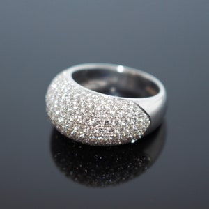 May include: A wide, silver-toned ring with a curved surface covered in small, sparkling clear stones. The ring has a smooth, polished band and a rounded, gem-studded top. The ring is displayed on a reflective black surface.