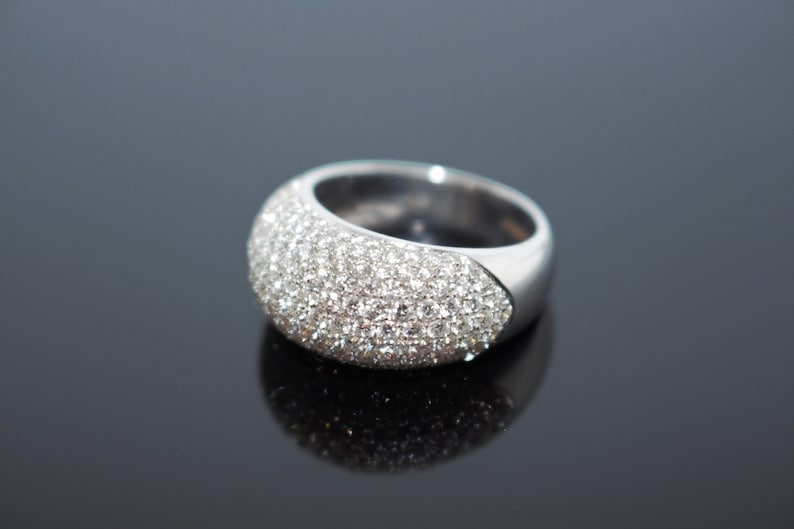 May include: A wide, silver-toned ring with a curved surface covered in small, sparkling clear stones. The ring has a smooth, polished band and a rounded, gem-studded top. The ring is displayed on a reflective black surface.