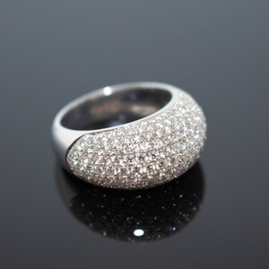 May include: A silver ring with a rounded top, fully encrusted with small, sparkling crystals. The ring sits on a reflective black surface, highlighting its brilliant facets. The band is smooth and polished.