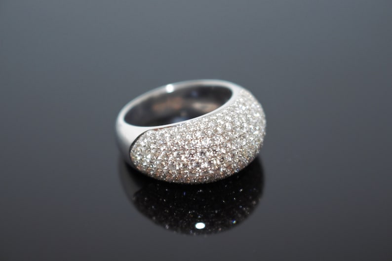 May include: A silver ring with a rounded top, fully encrusted with small, sparkling crystals. The ring sits on a reflective black surface, highlighting its brilliant facets. The band is smooth and polished.