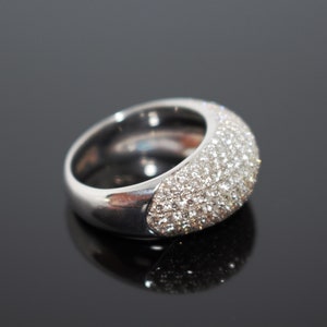 May include: A silver ring with a wide band and a domed top. The top of the ring is covered in small, sparkling stones, creating a dazzling effect. The ring is set against a dark, reflective surface.