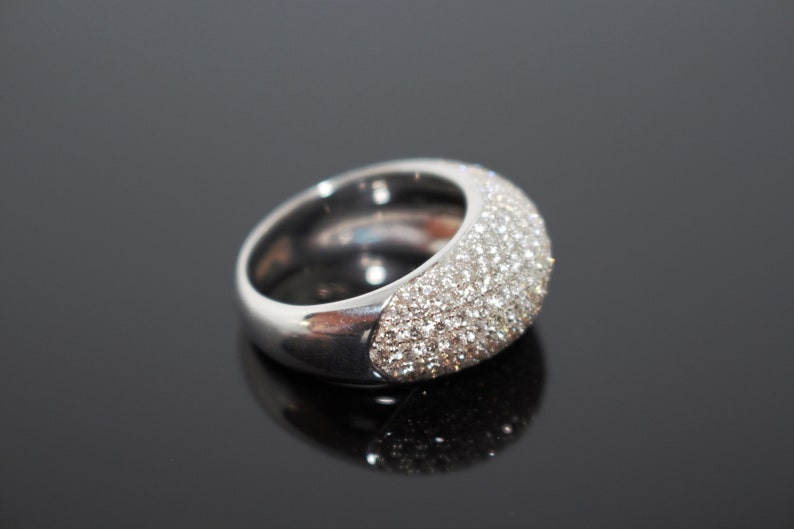 May include: A silver ring with a wide band and a domed top. The top of the ring is covered in small, sparkling stones, creating a dazzling effect. The ring is set against a dark, reflective surface.