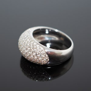 May include: A silver-toned ring with a rounded, wide band. The top half of the ring is encrusted with small, sparkling stones. The ring is set against a dark, reflective surface, highlighting its polished finish.
