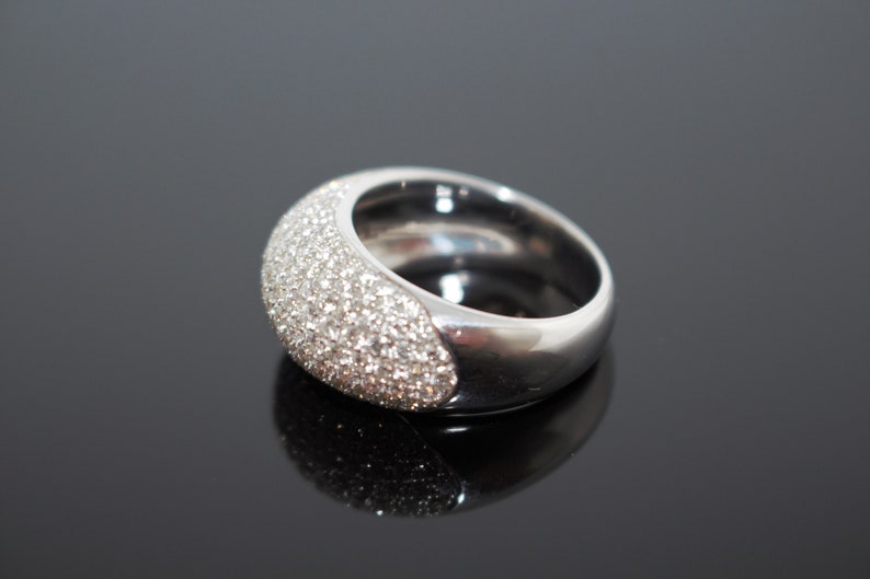 May include: A silver-toned ring with a rounded, wide band. The top half of the ring is encrusted with small, sparkling stones. The ring is set against a dark, reflective surface, highlighting its polished finish.