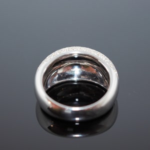 May include: A wide, silver-toned ring with a band of small, clear stones set along the top edge. The ring has a rounded, smooth design and reflects light, creating a sparkling effect. The ring is likely made of a precious metal.