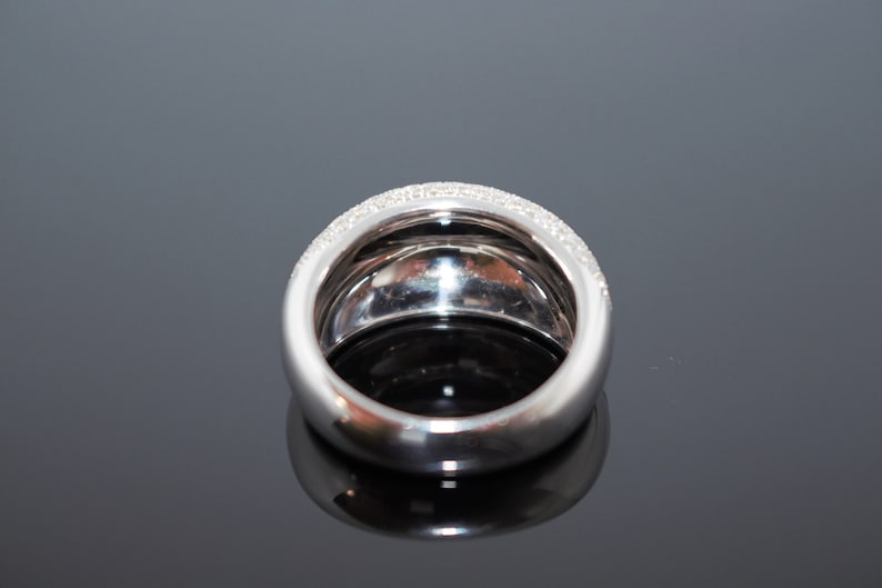 May include: A wide, silver-toned ring with a band of small, clear stones set along the top edge. The ring has a rounded, smooth design and reflects light, creating a sparkling effect. The ring is likely made of a precious metal.