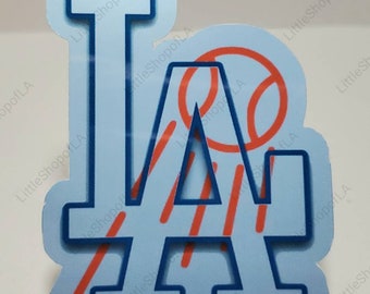 May include: A light blue sticker featuring the letters "LA" in a stylized design, with a baseball incorporated. The letters are outlined in dark blue, with red diagonal lines and a red baseball. The sticker has a white border.