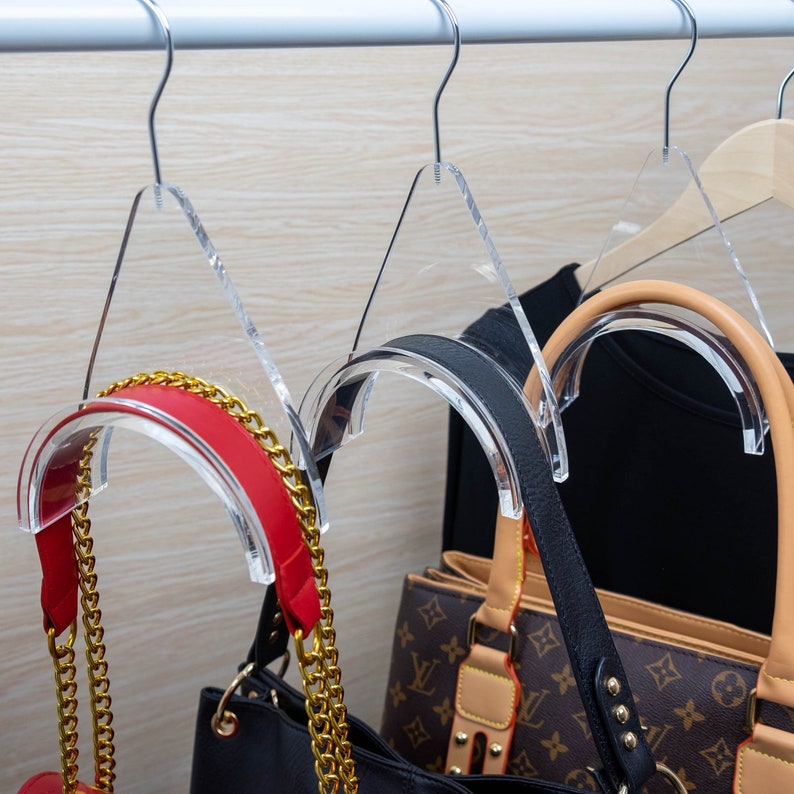 Puede incluir: Tres perchas de acr&iacute;lico transparente para bolsos de mano con un bolso de mano negro, marr&oacute;n y tostado colgado de ellas. Las perchas est&aacute;n dise&ntilde;adas para sujetar las correas de los bolsos de mano.