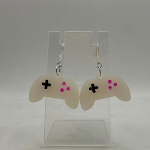 Puede incluir: Un par de pendientes de plástico blanco opaco con forma de mando de videojuego. El mando tiene botones negros y detalles en rosa.