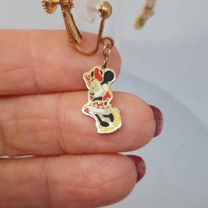 May include: A pair of gold-tone clip-on earrings featuring a red, white, and black Minnie Mouse charm. The charm is attached to the earring with a gold chain.