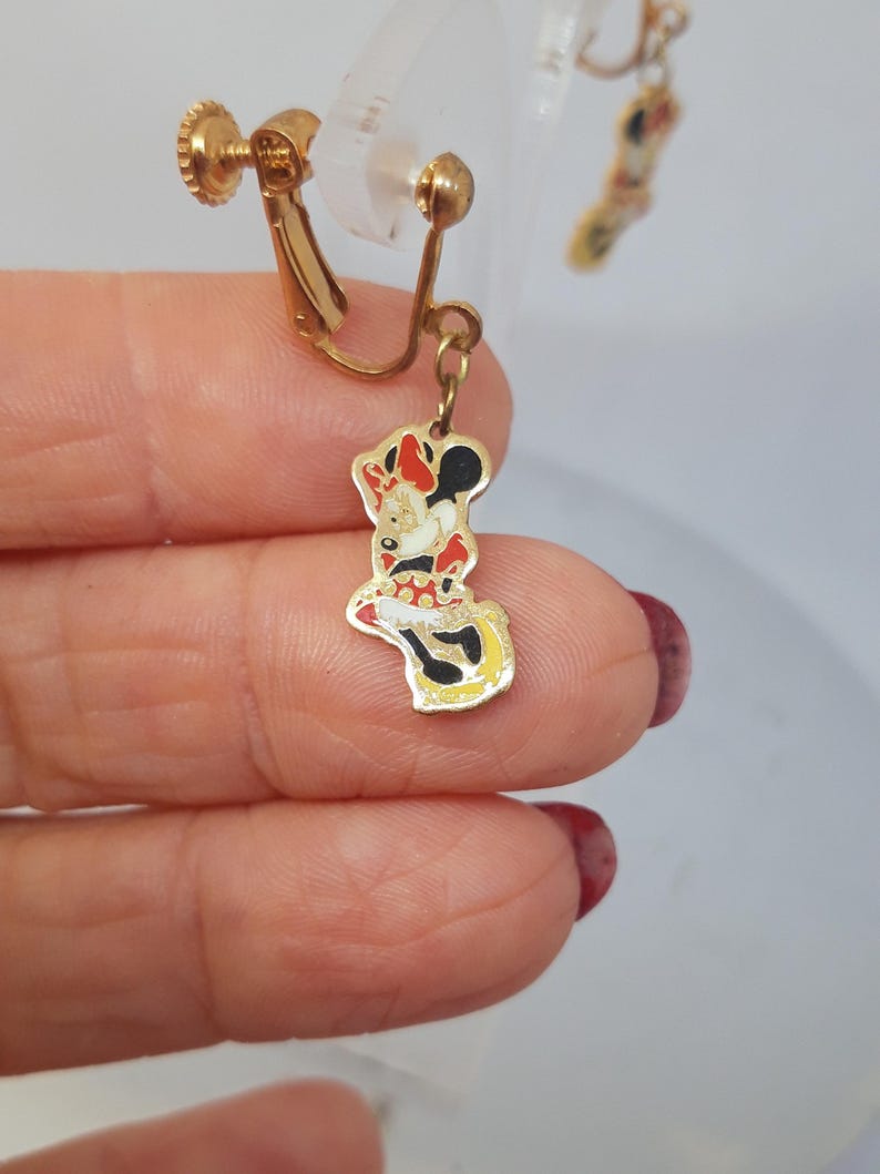 May include: A pair of gold-tone clip-on earrings featuring a red, white, and black Minnie Mouse charm. The charm is attached to the earring with a gold chain.