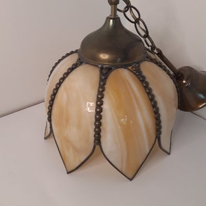 May include: A brass and glass pendant light fitting with a chain. The glass shade is a pale yellow with a white swirl pattern and has a scalloped edge. The shade is decorated with small brass beads.