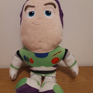 Może przedstawiać: Pluszowa zabawka Buzz Lightyear z serii Toy Story. Zabawka siedzi z wyciągniętymi rękami. Zabawka ma na sobie zielono-biały skafander kosmiczny z fioletowymi akcentami. Zabawka ma fioletowy hełm i białą twarz z haftowanymi cechami. Zabawka ma czerwoną i białą naszywkę na piersi z napisem "LIGHTYEAR".