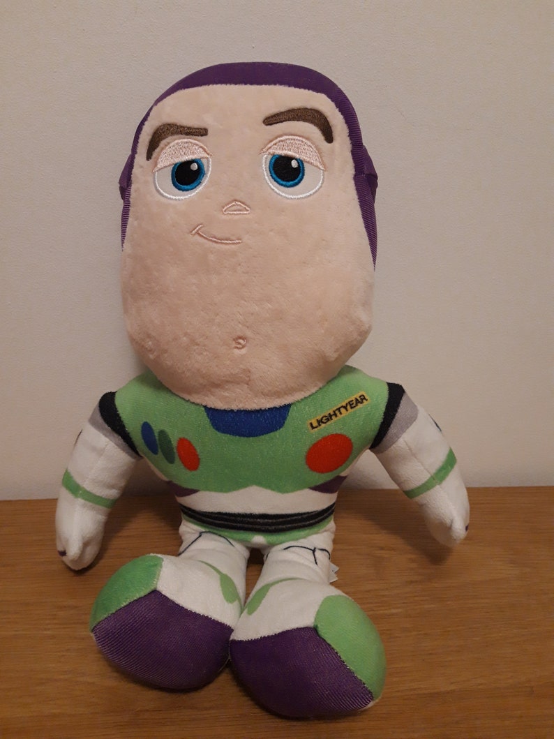 Może przedstawiać: Pluszowa zabawka Buzz Lightyear z serii Toy Story. Zabawka siedzi z wyciągniętymi rękami. Zabawka ma na sobie zielono-biały skafander kosmiczny z fioletowymi akcentami. Zabawka ma fioletowy hełm i białą twarz z haftowanymi cechami. Zabawka ma czerwoną i białą naszywkę na piersi z napisem "LIGHTYEAR".