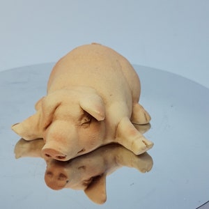 Puede incluir: Una figurita de cerdo de cerámica tumbada de espaldas con las patas extendidas. El cerdo es de color marrón claro y tiene una cara detallada.
