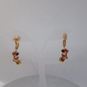 May include: A pair of gold-tone clip-on earrings featuring a cartoon image of Minnie Mouse in a red dress and yellow shoes.