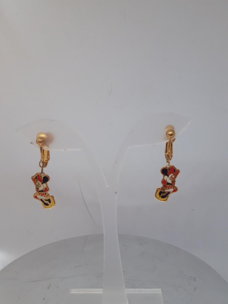 May include: A pair of gold-tone clip-on earrings featuring a cartoon image of Minnie Mouse in a red dress and yellow shoes.