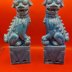 May include: A pair of glossy blue ceramic Foo Dog statues, each depicted in a seated position on a rectangular pedestal base. The figures feature detailed facial expressions with open mouths, stylised manes, and collars with hanging pendants.
