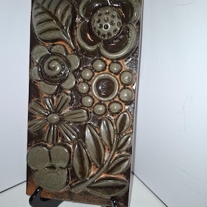 May include: A rectangular ceramic tile featuring a raised floral pattern. The flowers and leaves are in shades of green and brown, set against a dark brown background. The tile is displayed on a black stand.
