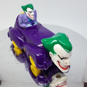 May include: A purple toy car with a Joker figure driving and another Joker figure attached to the front. The car has yellow wheels and the Joker figures have green hair and white faces with a wide grin.